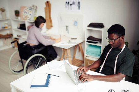 Téléchargez les photos : Créatif jeune créateur de mode noir travaillant sur ordinateur portable lorsque son collègue esquisse en arrière-plan - en image libre de droit