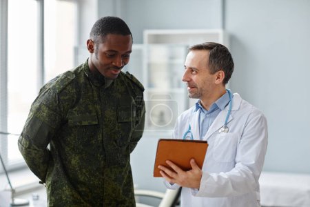 Photo for Mature Caucasian doctor in white coat showing good checkup results to young Black military officer pn digital tablet screen - Royalty Free Image