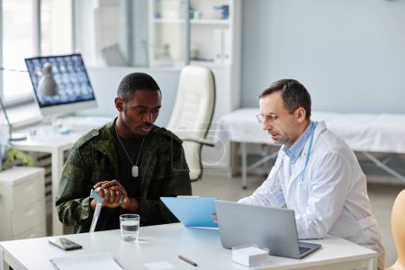Photo for Mature Caucasian physician showing medical papers on clipboard to young Black soldier and explaining something during consultation - Royalty Free Image