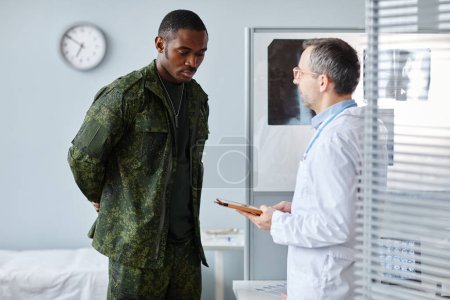 Photo for Mature Caucasian doctor holding digital tablet standing in front of young Black military man talking about treatment - Royalty Free Image