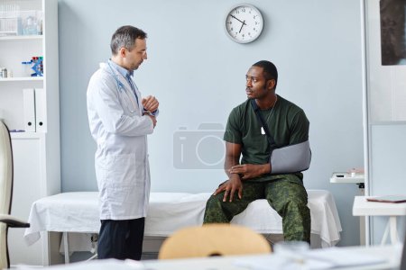 Photo for Young Black army officer wearing arm sling sitting on medical couch talking to mature Caucasian doctor - Royalty Free Image