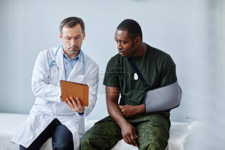Photo for Mature traumatologist sitting on medical couch with young Black military officer showing him arm x-ray on digital tablet - Royalty Free Image
