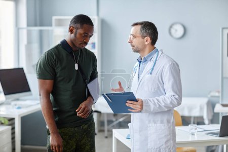 Photo for Mature traumatologist holding clipboard discussing something with young Black soldier with arm injured - Royalty Free Image