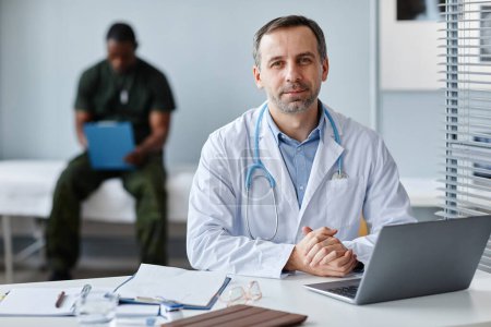 Téléchargez les photos : Portrait d'un médecin caucasien mature moderne travaillant dans un hôpital militaire, son patient assis sur le canapé remplissant un formulaire de contrôle médical sur fond - en image libre de droit