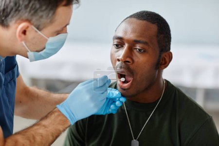 Foto de Médico maduro irreconocible usando máscara y guantes tomando la prueba de hisopo bucal de un joven oficial del ejército afroamericano - Imagen libre de derechos