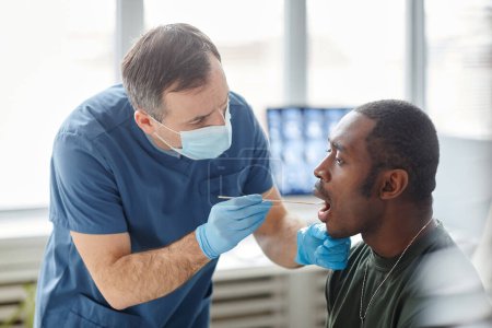 Téléchargez les photos : Travailleur médical portant un masque protecteur et des gants prenant écouvillonnage de la bouche du jeune soldat noir pour le test PCR - en image libre de droit