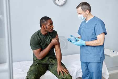 Téléchargez les photos : Infirmière masculine mature donnant des recommandations au jeune soldat noir après lui avoir administré une injection de vaccin contre le coronavirus - en image libre de droit