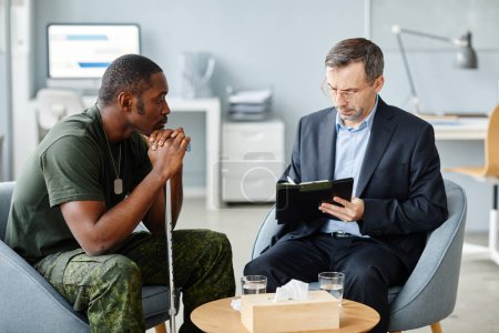 Photo for Sad young adult African American war veteran with walking stick sitting on chair watching his psychotherapist making notes during session - Royalty Free Image