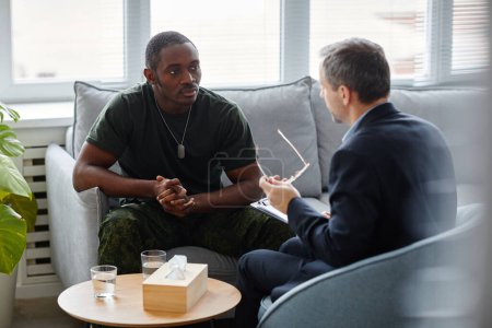 Téléchargez les photos : Jeune Afro-Américain en uniforme militaire assis sur le canapé à écouter un psychothérapeute pendant une séance de thérapie - en image libre de droit