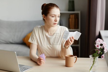 Téléchargez les photos : Portrait de jeune femme blanche lisant le manuel d'instruction pour la coupe menstruelle à la maison dans l'intérieur minimal, espace de copie - en image libre de droit
