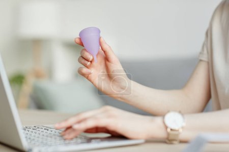 Side view closeup of young woman holding pink menstrual cup by laptop and doing research on feminine hygiene products
