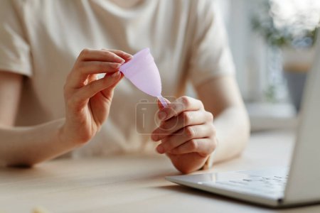 Téléchargez les photos : Gros plan de la jeune femme tenant tasse menstruelle rose par ordinateur portable et la recherche de produits féminins - en image libre de droit