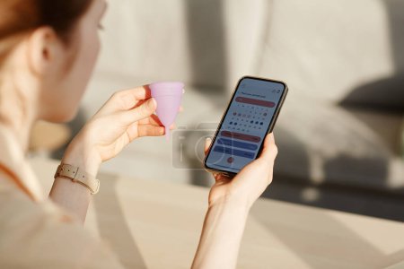 Téléchargez les photos : Gros plan de la jeune femme calculant le cycle menstruel à l'aide d'une application mobile et tenant une tasse réutilisable - en image libre de droit