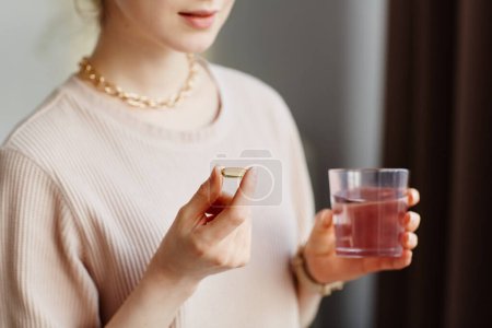 Téléchargez les photos : Gros plan minimal de la jeune femme tenant une capsule vitaminique tout en prenant des suppléments avec un verre d'eau à la maison - en image libre de droit