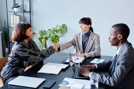 Téléchargez les photos : Équipe d'affaires moderne serrant la main à la table de réunion dans le bureau se saluant et souriant - en image libre de droit
