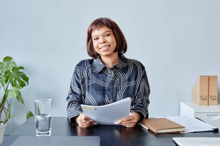 Foto de Retrato mínimo de una joven negra con corte de pelo corto leyendo documentos en el lugar de trabajo en la oficina y sonriendo a la cámara - Imagen libre de derechos
