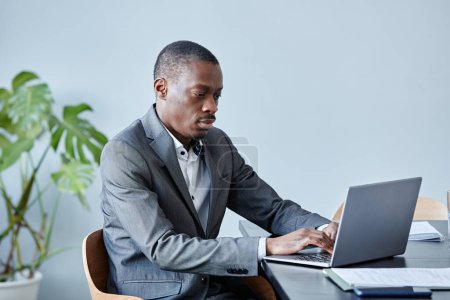 Photo for Minimal portrait of professional black executive using laptop while sitting at workplace against blue wall in office and wearing suit, copy space - Royalty Free Image