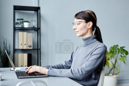 Téléchargez les photos : Vue latérale minime portrait de jeune femme d'affaires portant des lunettes et souriant tout en utilisant un ordinateur portable dans le bureau contre le bleu - en image libre de droit