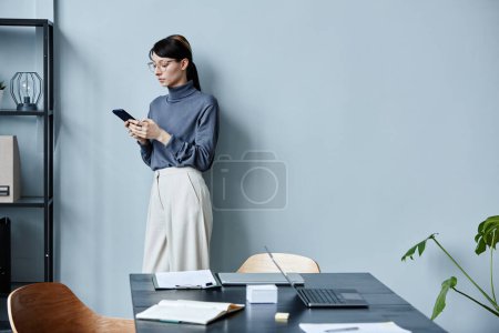 Photo for Minimal portrait of young businesswoman holding smartphone while leaning against pale blue wall and wearing casual outfit in office, copy space - Royalty Free Image