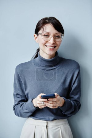 Téléchargez les photos : Portrait franc de jeune femme d'affaires tenant smartphone et souriant à la caméra sur fond bleu simple - en image libre de droit