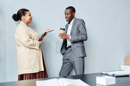 Téléchargez les photos : Vue latérale minime portrait de deux hommes d'affaires afro-américains bavardant occasionnellement tout en s'appuyant contre le mur bleu dans le bureau, espace de copie - en image libre de droit