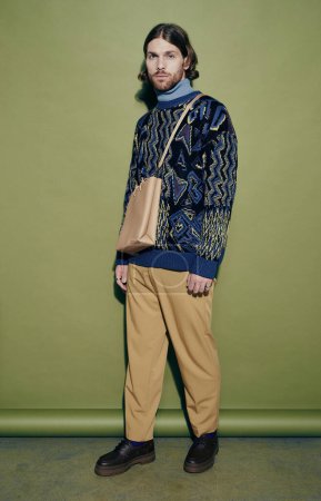 Foto de Tirador vertical de cuerpo entero de modelo de moda masculino con suéter de moda y bolso bandolera sobre fondo verde - Imagen libre de derechos
