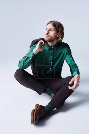Foto de Retrato de tendencia del modelo de moda masculino posando casualmente usando traje con camisa de seda verde azulado y sentado en el suelo - Imagen libre de derechos