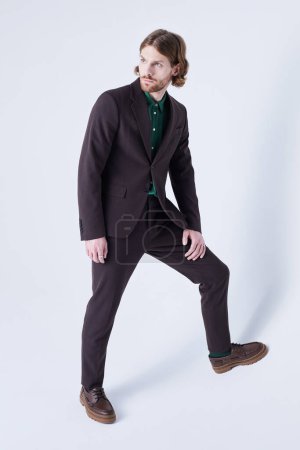 Foto de Mínimo tiro de longitud completa de modelo de moda masculina con traje en tono terroso y mirando hacia otro lado contra el gris - Imagen libre de derechos