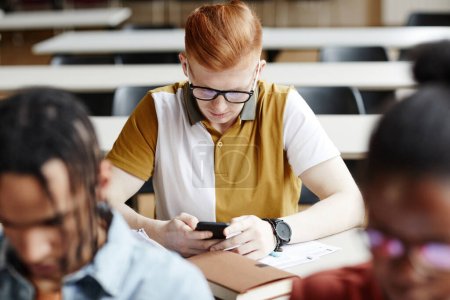 Téléchargez les photos : Portrait grand angle de jeune homme roux utilisant un smartphone au bureau dans une salle de classe universitaire - en image libre de droit