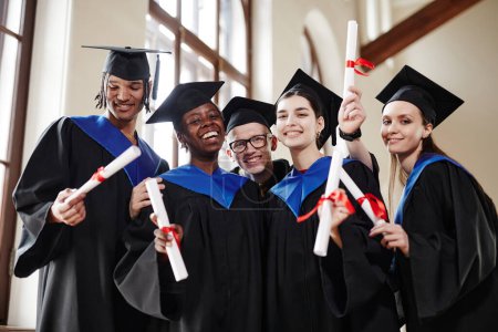 Photo for Group of joyful young people wearing graduation gowns at ceremony in university and smiling at camera - Royalty Free Image