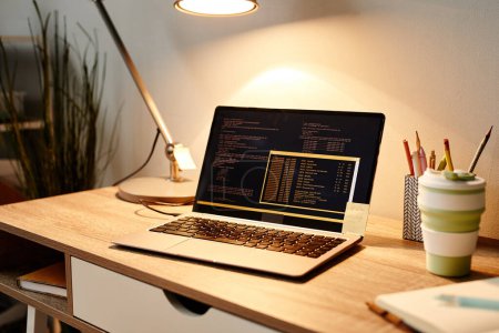 Foto de Primer plano de la computadora portátil con código en la pantalla en el acogedor lugar de trabajo de la oficina en casa iluminado por la luz de la lámpara, espacio de copia - Imagen libre de derechos