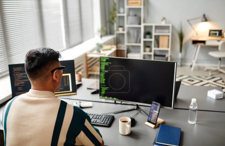 Téléchargez les photos : Vue arrière du développeur informatique masculin tapant sur le clavier avec le code de programmation sur l'écran de l'ordinateur tout en travaillant dans l'intérieur du bureau, espace de copie - en image libre de droit
