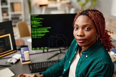 Téléchargez les photos : Portrait de jeune développeuse informatique regardant la caméra contre le code de programmation sur l'écran d'ordinateur à l'intérieur du bureau - en image libre de droit