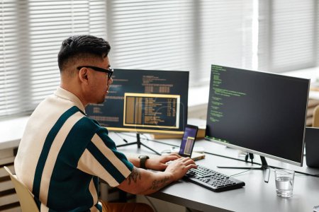 Side view of Asian IT developer typing on keyboard with programming code on computer screen while working in office
