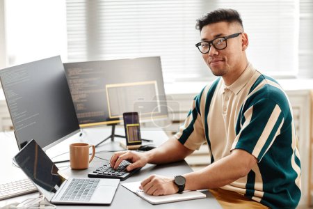 Téléchargez les photos : Portrait chaleureux de développeur informatique asiatique ou ingénieur QA regardant la caméra tout en étant assis sur le lieu de travail avec des ordinateurs, copier l'espace - en image libre de droit