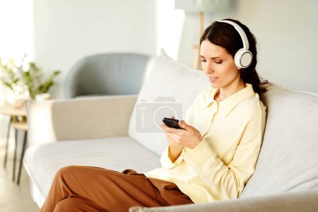 Photo for Young adult Caucasian woman wearing headphones sitting on couch at home choosing music to listen to on smartphone - Royalty Free Image