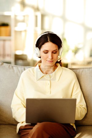 Photo for Vertical shot of beautiful young adult Caucasian woman wearing casual clothes and headphones sitting on sofa at home working remotely using laptop - Royalty Free Image