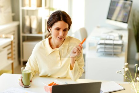 Photo for Modern young adult Caucasian woman sitting at table in office having fresh carrots and smoothie for snack and talking on video call with co-worker - Royalty Free Image
