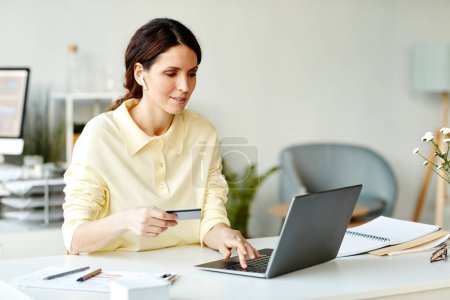 Foto de Horizontal media toma de joven mujer caucásica adulto sentado en el escritorio en la oficina usando el ordenador portátil y la tarjeta bancaria para pedir comida a domicilio - Imagen libre de derechos
