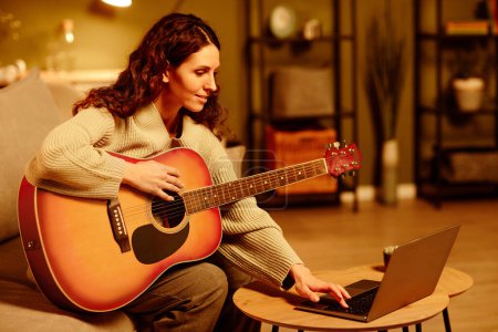 Photo for Portrait of young adult Caucasian woman learning to play guitar watching lessons in Internet on laptop in evening - Royalty Free Image