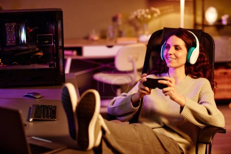 Foto de Moderno adulto joven mujer caucásica con ropa casual y auriculares pasar la noche jugando videojuegos con el uso de controlador - Imagen libre de derechos