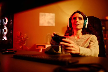Téléchargez les photos : Jeune femme blanche adulte avec de longs cheveux bouclés portant des écouteurs profiter de jouer à un jeu vidéo la nuit - en image libre de droit