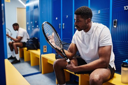 Téléchargez les photos : Portrait d'un sportif noir musclé tenant une raquette de tennis tout en se préparant à l'entraînement dans un vestiaire, espace de copie - en image libre de droit