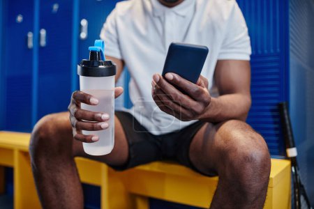 Téléchargez les photos : Gros plan d'un sportif noir musclé utilisant un smartphone dans un vestiaire et tenant une bouteille d'eau - en image libre de droit