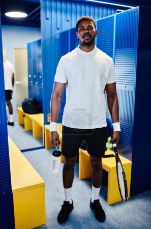 Téléchargez les photos : Portrait vertical complet du joueur de tennis noir regardant la caméra tout en se tenant dans les vestiaires - en image libre de droit