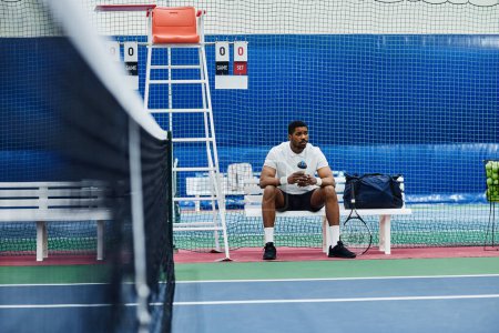 Photo for Wide angle shot of professional black sportsman sitting on bench in tennis court and holding water bottle, copy space - Royalty Free Image
