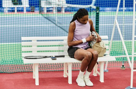 Téléchargez les photos : Portrait complet de la jeune joueuse de tennis féminine se préparant pour la pratique tout en étant assise sur un banc au court de tennis, espace de copie - en image libre de droit