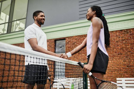 Téléchargez les photos : Vue latérale de deux joueurs de tennis souriants serrant la main à travers le filet pendant le match au court - en image libre de droit