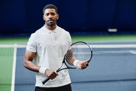 Photo for Waist up portrait of African American tennis player looking at camera while posing confidently with racket at indoor court, copy space - Royalty Free Image