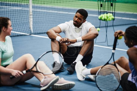 Téléchargez les photos : Groupe de personnes se détendre au court de tennis intérieur et bavarder tout en étant assis sur le sol - en image libre de droit